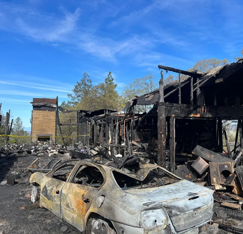 A burned wood-frame house and a fire-damaged car after a wildfire.
