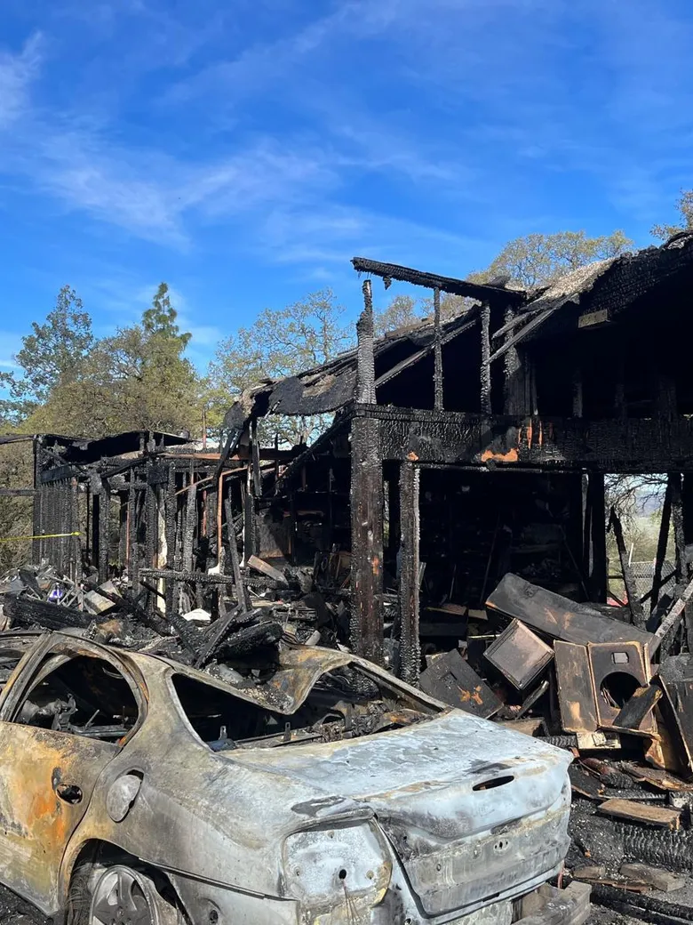 A wood frame house and a car burned out after a fire, the brick chimney still standing.