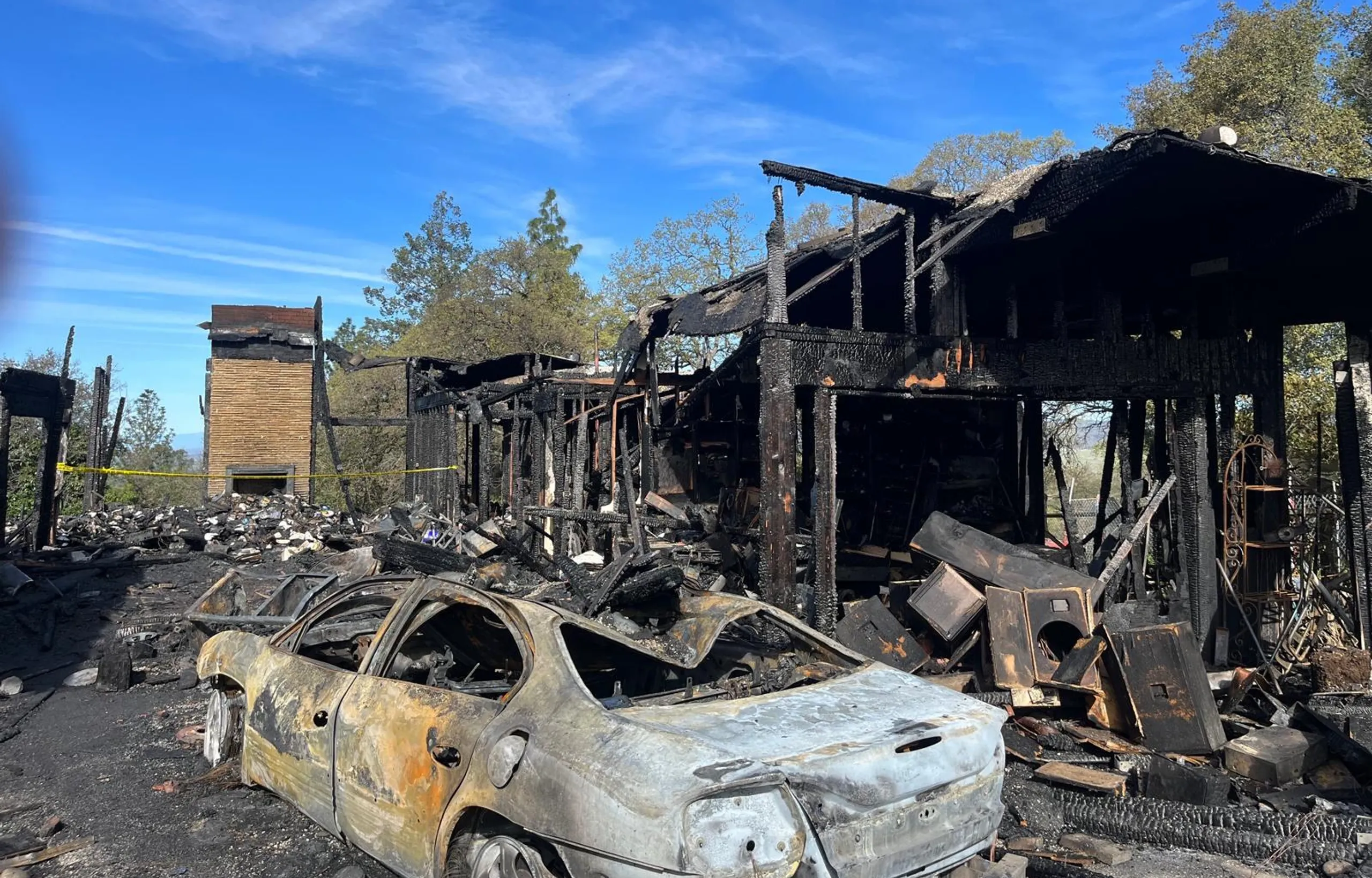 A wood frame house and a car burned out after a fire, the brick chimney still standing.